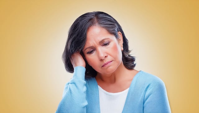 A woman with short, dark hair looks down thoughtfully, resting her head on her right hand. She wears a light blue sweater over a white shirt.