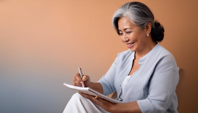 A mature Asian woman with silver hair styled in a bun smiles warmly while writing in a notebook. She wears a light blue cardigan and white dress, sitting against a peachy-orange backdrop.