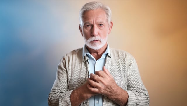 An older man with white hair and a beard stands with his hands clasped at chest level. He wears a light-colored, patterned shirt over a blue button-up. The background transitions smoothly from blue to orange.