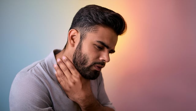 A man with a neatly groomed beard and short hair touches his neck with one hand, appearing in discomfort. He wears a textured light gray shirt, and his expression shows concern.