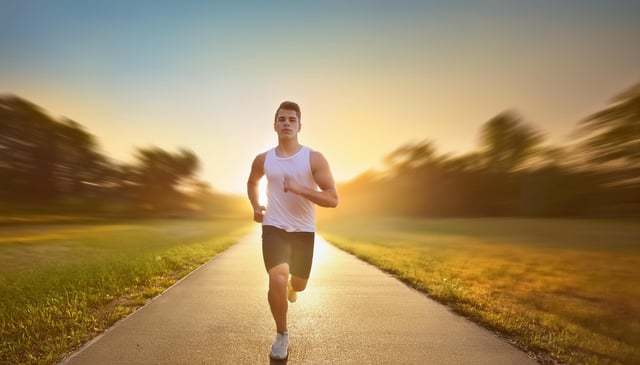 A person in a white tank top and black shorts runs energetically on a paved path. The setting sun casts a warm glow, highlighting their focused expression.