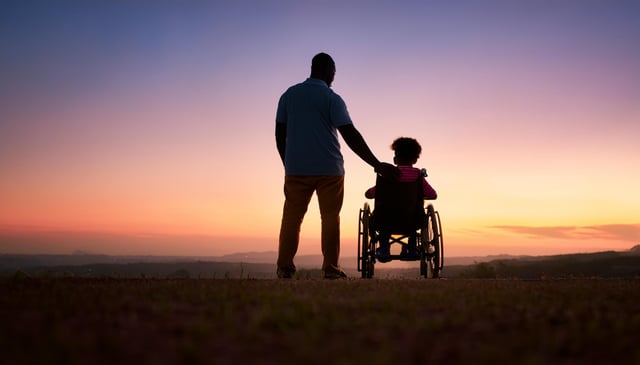 A man stands next to a child in a wheelchair, gently placing his hand on the child's shoulder. The pair faces a colorful sunset, silhouetted against the sky.