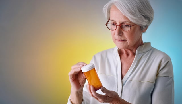 An elderly woman with short white hair, wearing red glasses and a light-colored blouse, examines an orange prescription bottle intently with both hands. Her expression appears focused and concerned.