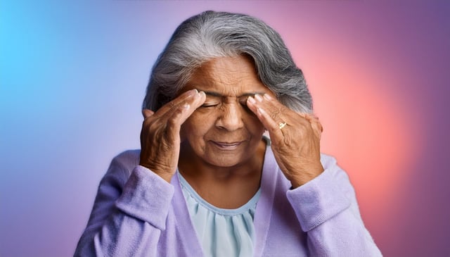 An elderly woman with gray hair presses her temples with both hands, eyes closed, wearing a lavender cardigan over a light blue shirt. She appears to be experiencing discomfort.