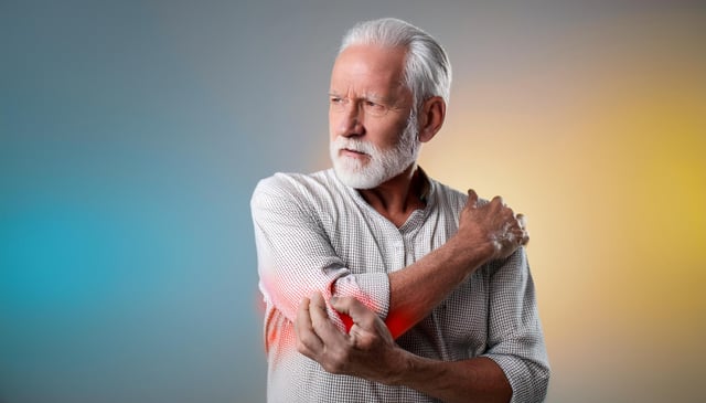 An elderly man with a white beard and hair grips his left elbow with his right hand. His expression conveys pain, and a reddish hue highlights his left forearm.