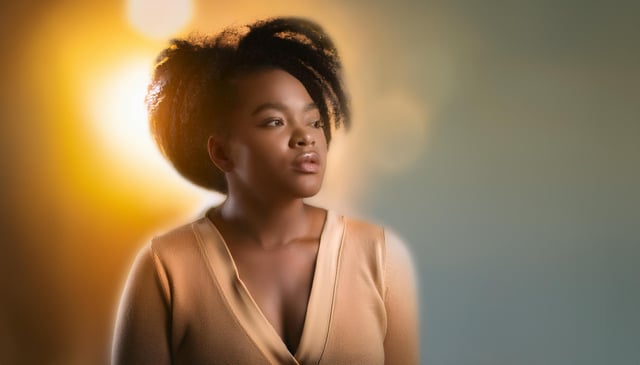 A woman gazes to the side with a thoughtful expression. She wears a beige top, and soft lighting highlights her hair, creating a warm, golden glow.