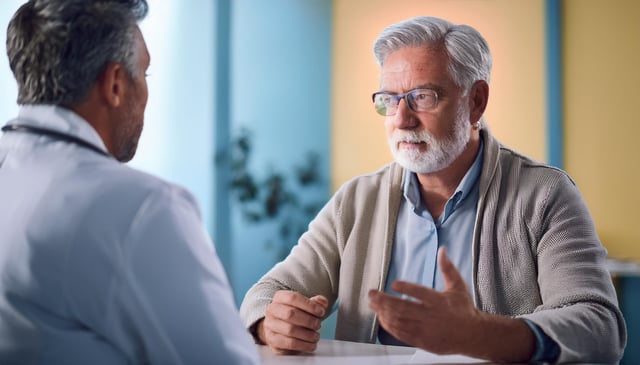 An older man with gray hair and a beard gestures while talking to another man, who wears a white coat and stethoscope. The conversation appears engaged and serious.