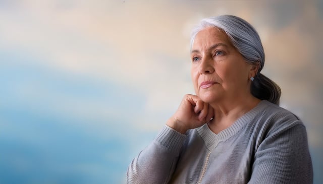 An elderly woman with long, gray hair tied back gazes thoughtfully to the side, resting her chin on her hand. She wears a gray sweater and stud earrings, exuding calmness.