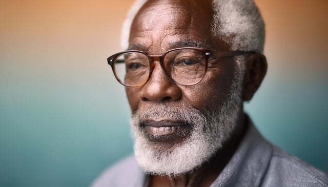 An older man with a white beard wears round glasses and a gentle expression. His serene face is highlighted against a soft gradient background, transitioning from orange to teal.