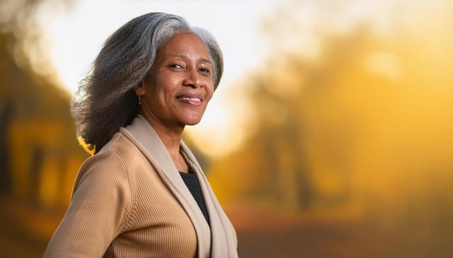 A woman with silver hair smiles warmly, wearing a beige cardigan over a dark top. The soft, golden light highlights her confident expression.