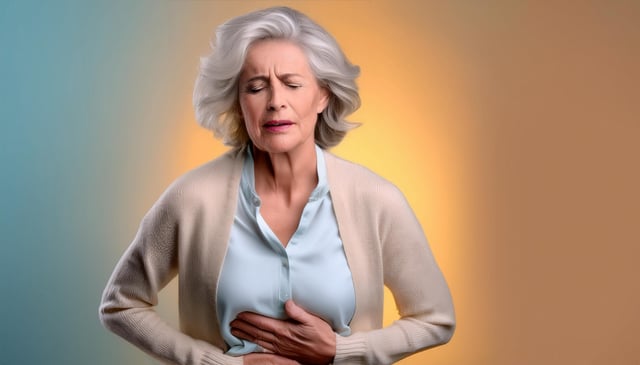 An older woman with white hair closes her eyes and presses her hands against her stomach, appearing in discomfort. She wears a beige cardigan over a light blue blouse.