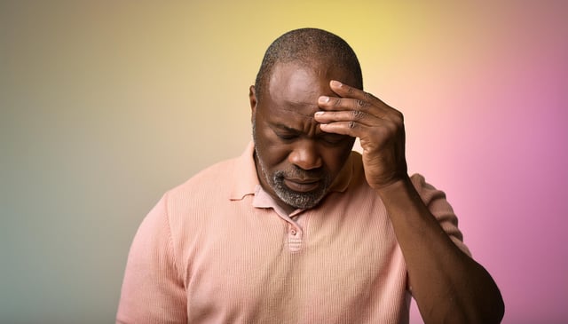 A man in a light pink polo shirt holds his forehead with his right hand, appearing to be in deep thought or distress. His eyes are closed, and his head is slightly bowed.