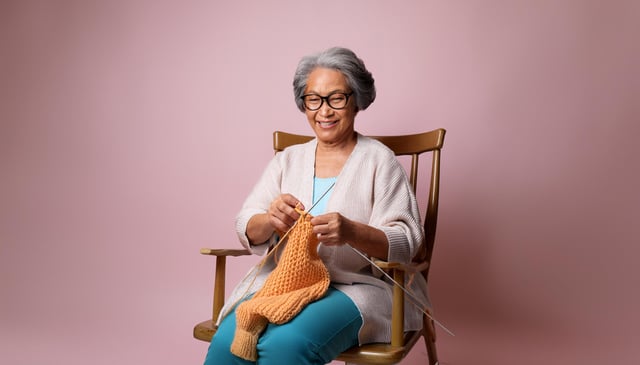 An older woman with short gray hair and glasses happily knits an orange scarf while sitting in a wooden rocking chair. She wears a light cardigan over a blue shirt and blue pants.