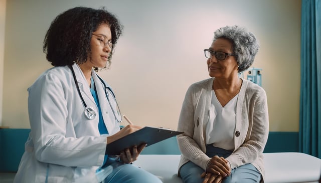 A doctor in a white coat and stethoscope consults with an elderly woman wearing glasses and a beige cardigan on an examination table. The doctor writes on a clipboard.