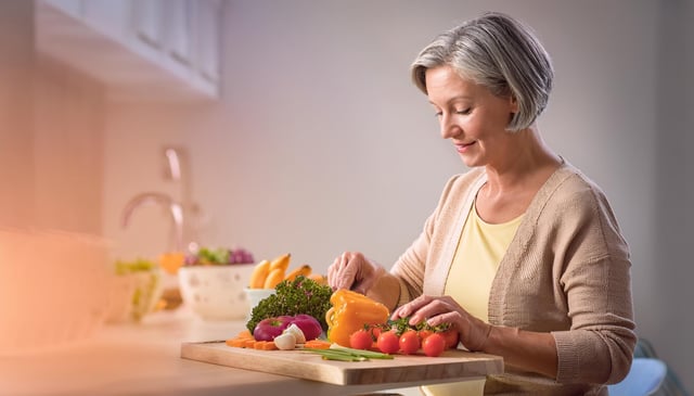 A woman slices vibrant vegetables, including a yellow bell pepper and vine tomatoes, on a wooden cutting board. She wears a beige cardigan and a yellow shirt, looking content and focused.