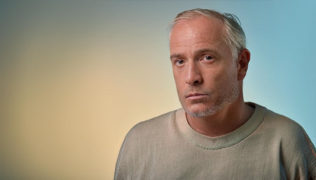 A middle-aged man with short gray hair and stubble gazes forward, wearing a beige sweater. Subtle lighting highlights his facial features against a soft gradient background.