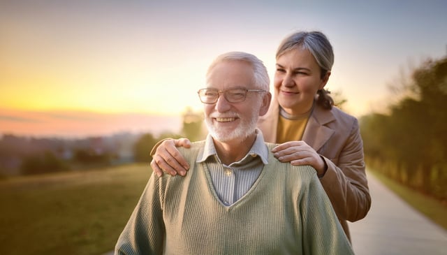 A smiling elderly man, wearing glasses and a green sweater, sits in the foreground. A woman stands behind him, gently resting her hands on his shoulders, smiling softly.